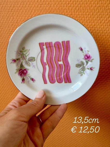 Witte schotel met de tekst "FUCK" in roze letters en bloemenrand. Prijs en maat vermeld.